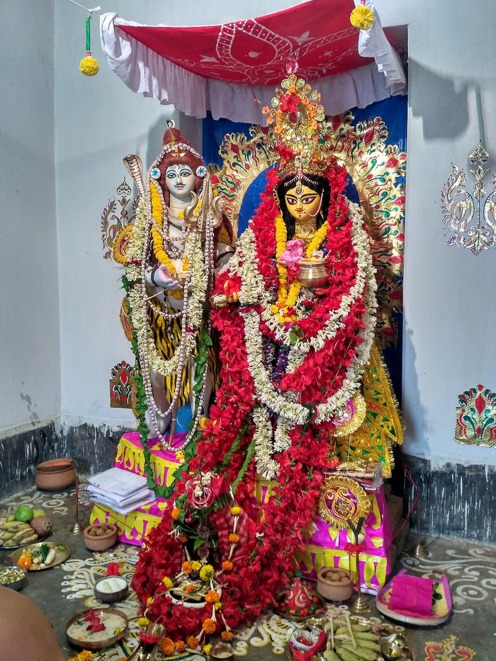 Garlanded Annapurna image beside Shiva, holding a food vessel, shown with ritual offerings in a worship shrine (2017).