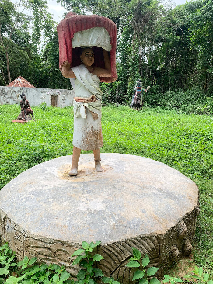 Arugba Ọya statue in Oya Grove: a votive-bearer carrying a covered vessel on her head, standing on a carved stone base.