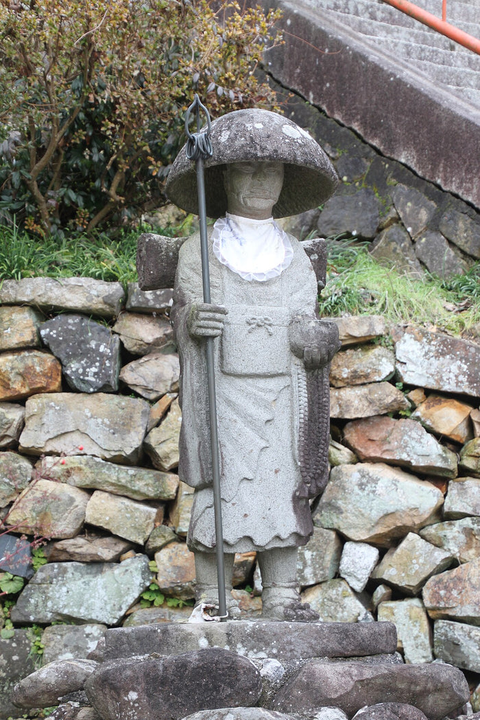 Weathered stone Daishi statue in pilgrim dress with hat and staff, standing by a temple stairway.