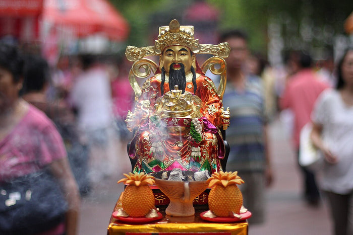 Small gilded statue of Caishen with a long black beard, shown as a household offering with incense and pineapples.