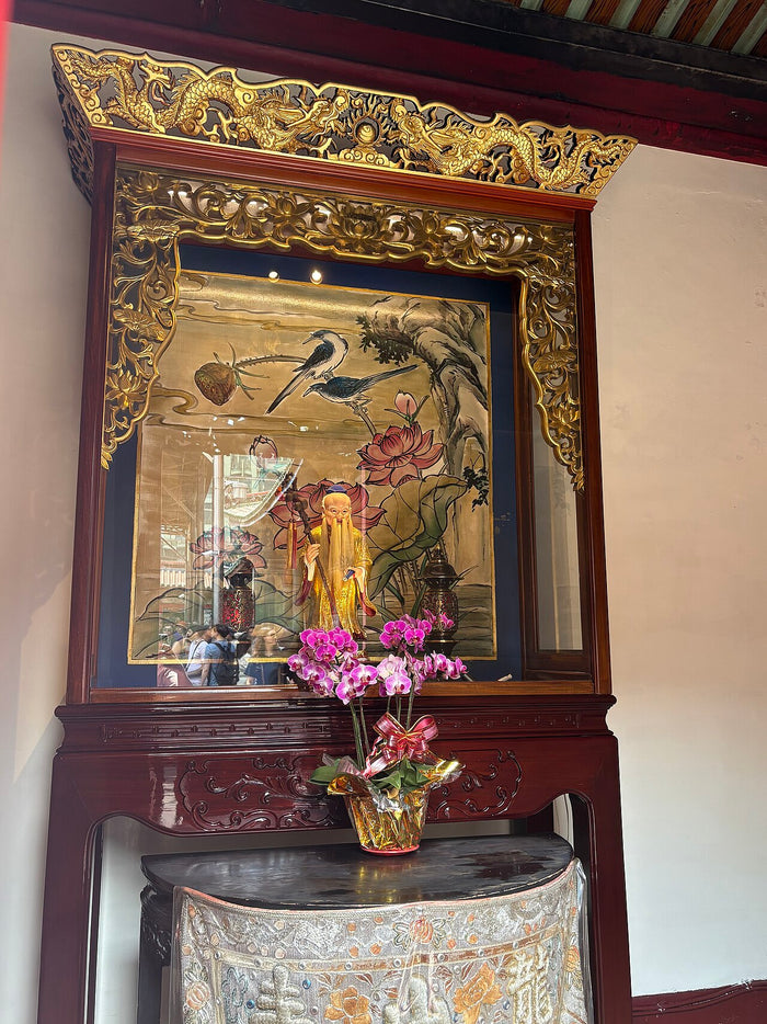 Yue Lao's shrine at Bangka Longshan Temple: a carved gilt-and-wood niche with a painted lotus-and-birds backdrop and an orchid offering.