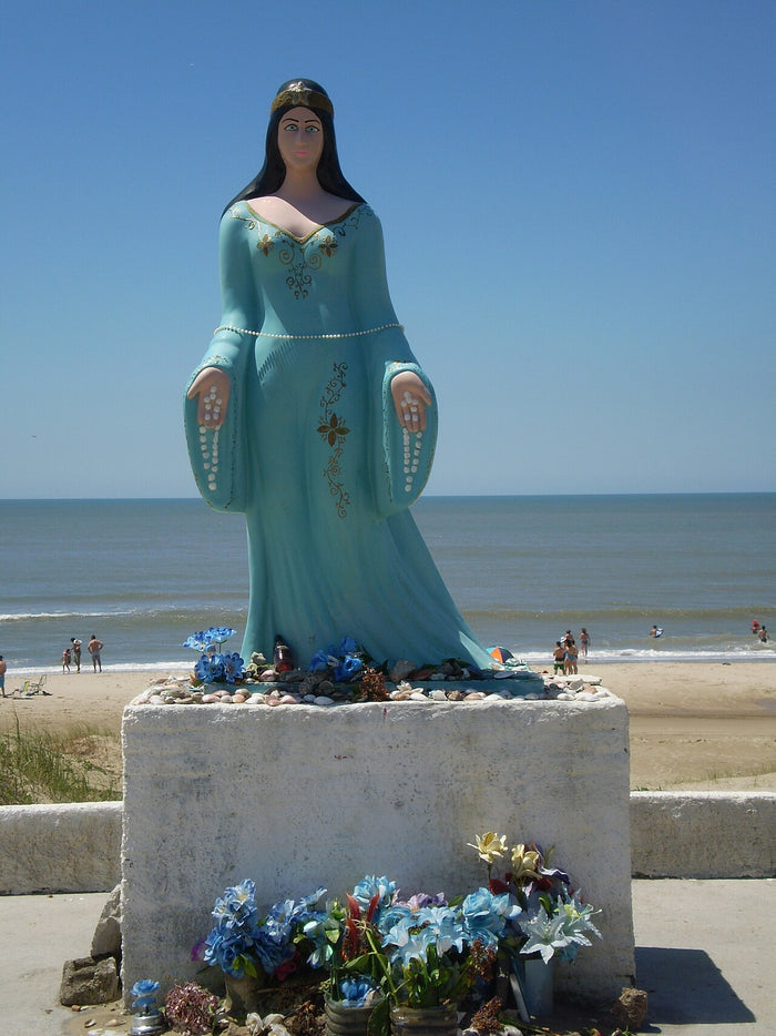 Iemanjá statue in blue-green robes on a seaside plinth, with blue flowers and shell offerings, in the familiar Brazilian image.