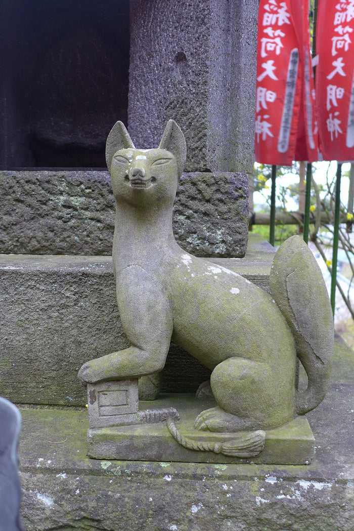 Stone Inari fox (kitsune) statue at a shrine, with its paw on a small pedestal, beside red Inari banners.