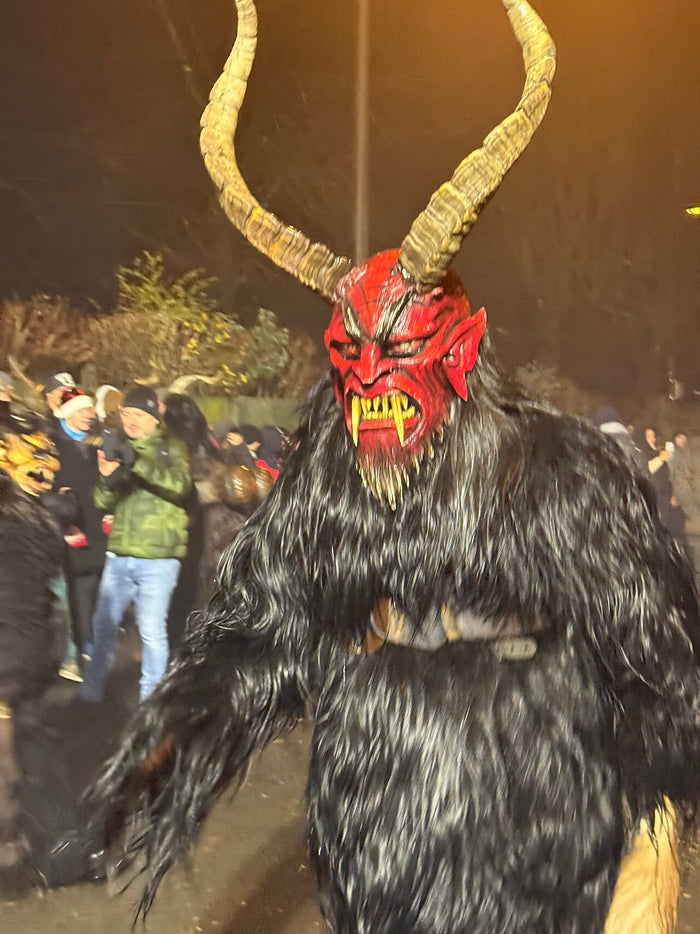 Krampus runner in black fur with long curved horns and a fanged red mask, at a December 2024 procession.