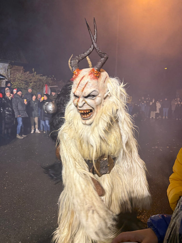 Krampus in white long-haired fur with a grimacing mask and dark horns, amid smoke at a December 2024 Krampus run.