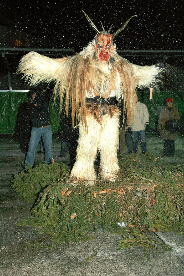 Krampus figure in shaggy fur with curved horns and red carved mask, standing on fir branches during a winter event.