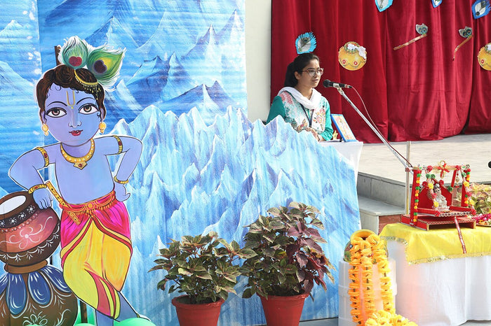 Painted school backdrop of child Krishna with peacock feather and butter pot, used in Janmashtami celebrations.