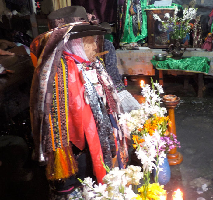 Maximón in Chichicastenango, wrapped in many scarves under a stacked hat, with flowers and candlelight in the shrine.