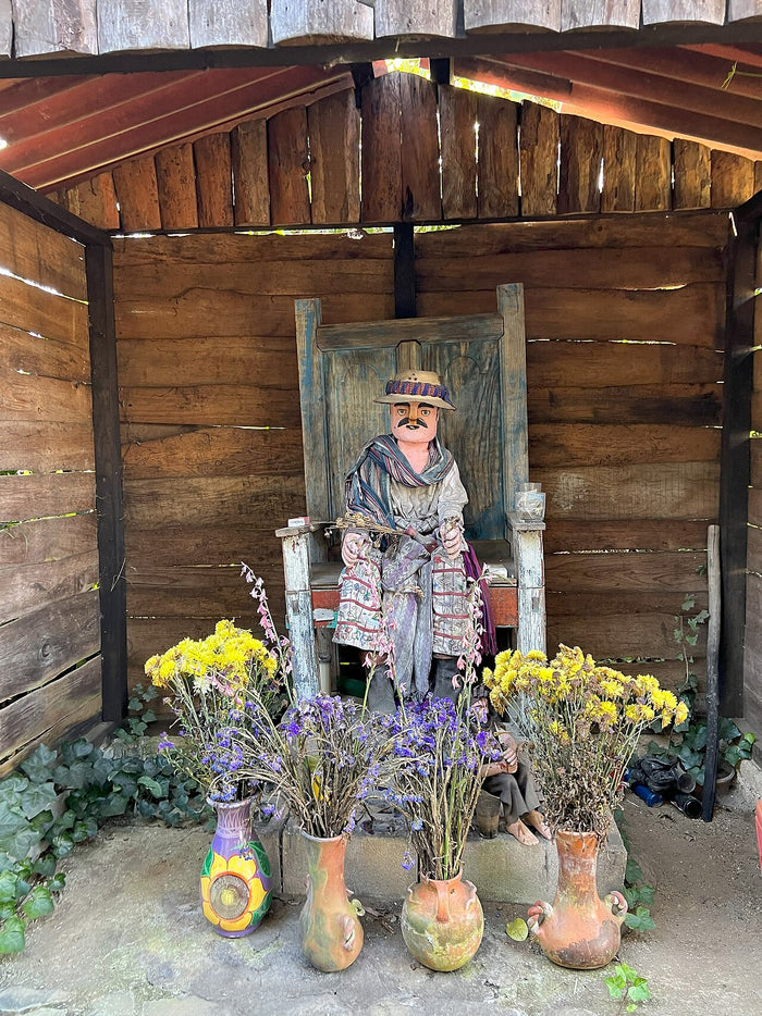 Roadside Maximón shrine in Antigua Guatemala (2023): a moustached figure on a wooden chair, with vases of flowers.