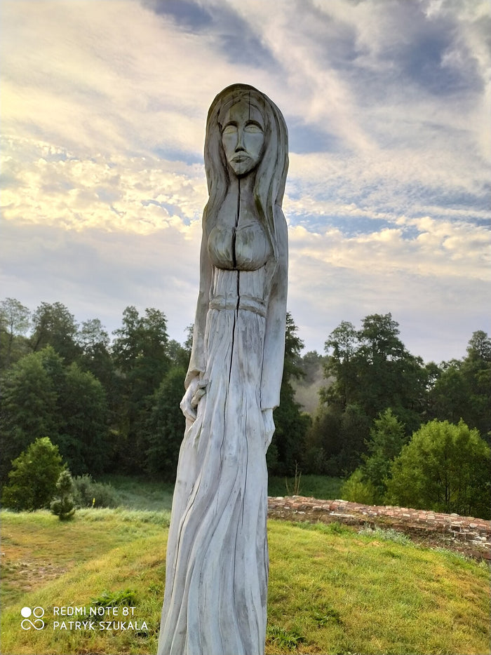 Modern wooden statue of Mokosz, a long-haired female figure, standing beside old earthworks. It shows her present-day veneration in Slavic Native Faith.