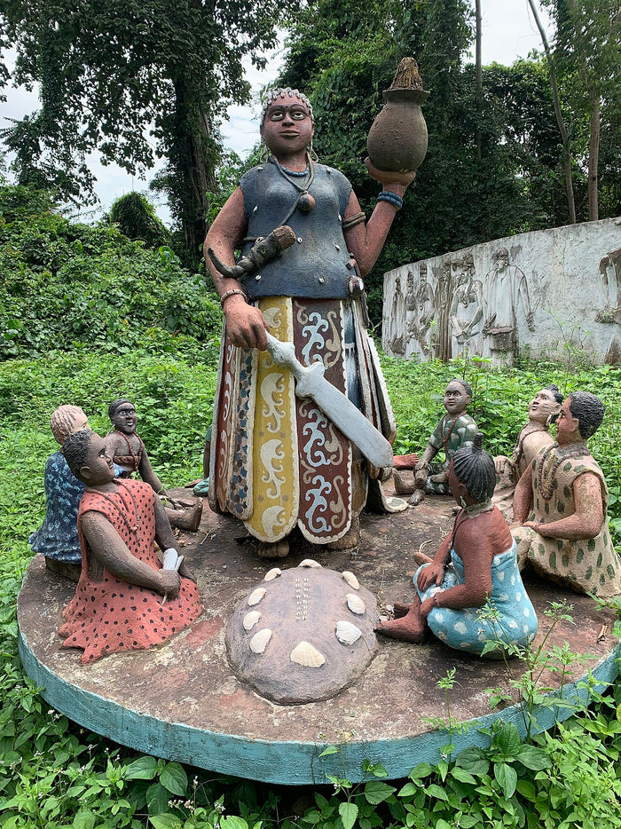 Statue of Ọya in Oya Grove holding a sword and a pot, with seated devotees around her.