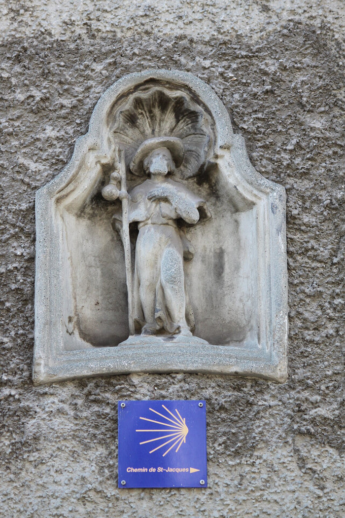 Small stone statue of Saint James as a pilgrim with staff, gourd, hat and scallop shell, above a Chemin de St-Jacques waymarker.