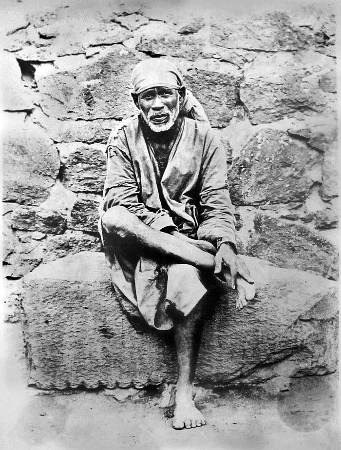 Shirdi Sai Baba seated cross-legged on a stone slab, c. 1915, wearing a cloth head-wrap and simple robe.