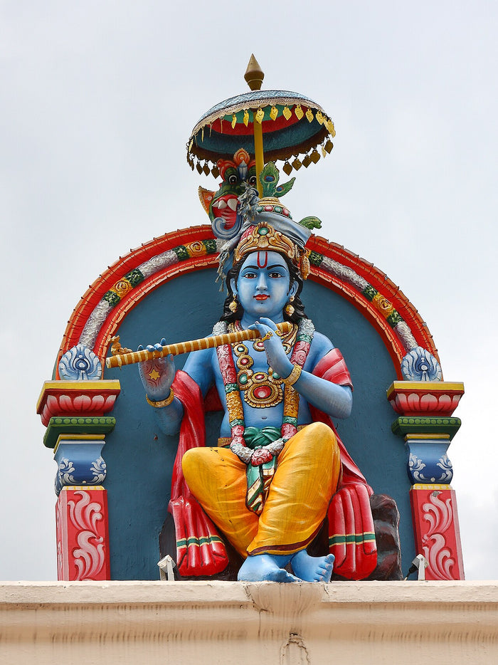 Painted temple sculpture of blue-skinned Krishna playing his flute beneath a parasol, a classic iconographic form.
