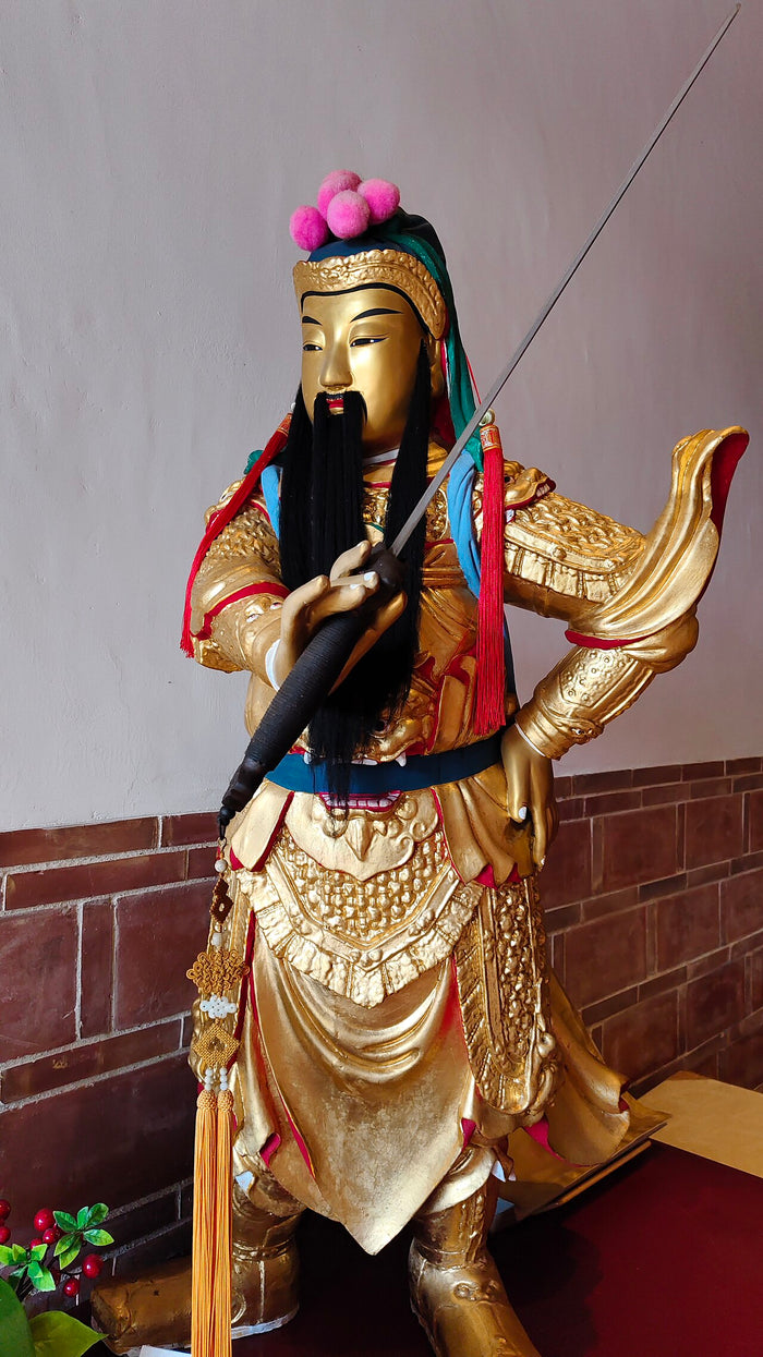 Standing gilded statue of Guan Gong in armor with long black beard, holding a sword.
