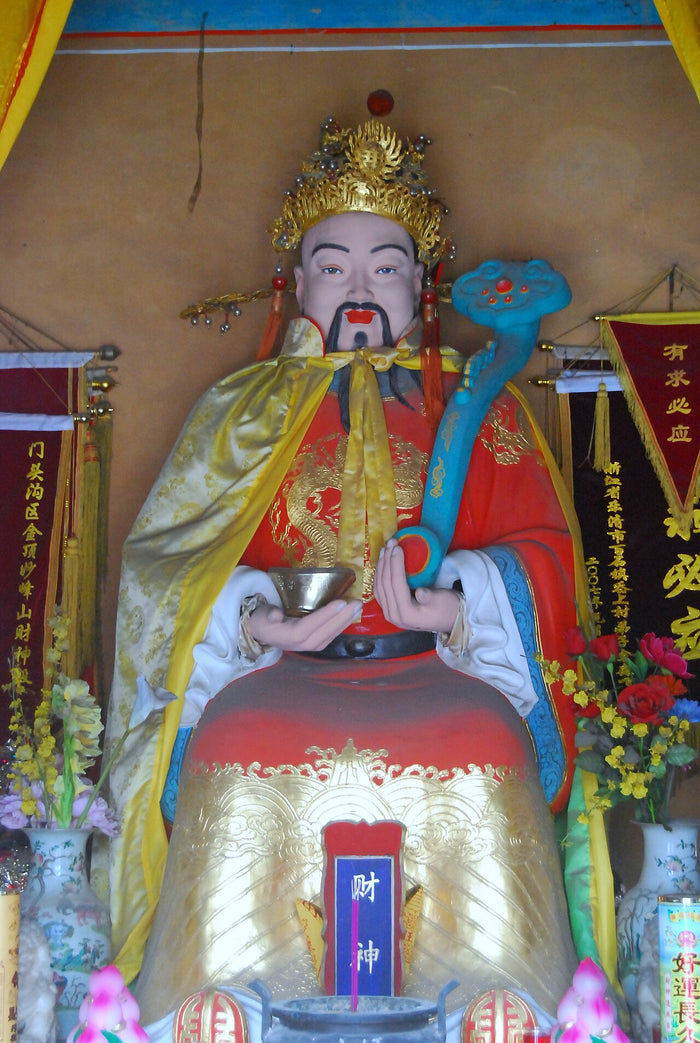 Painted statue of Caishen in a temple shrine, wearing a golden crown and red robes, holding a ruyi scepter and gold ingot.