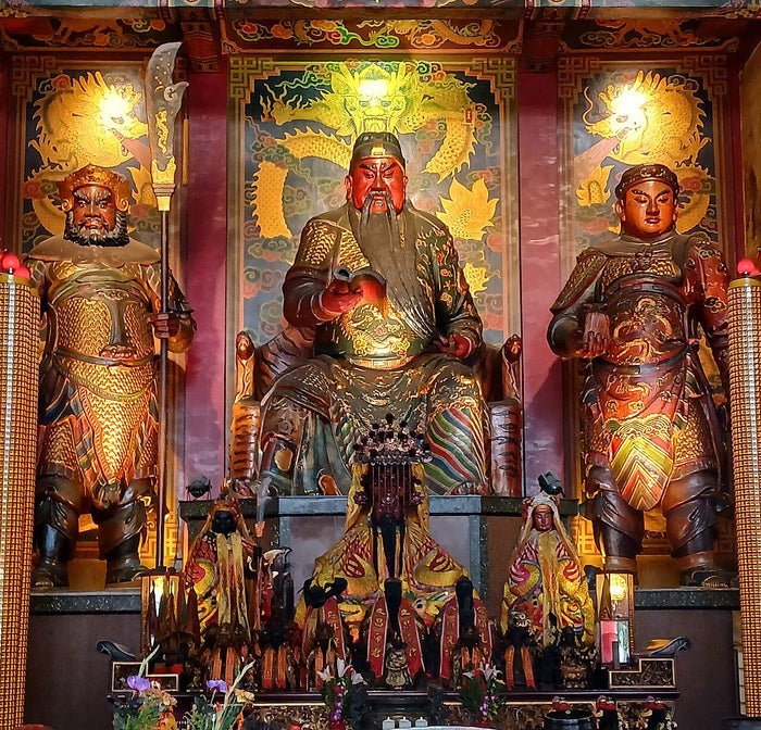 Enthroned red-faced Guan Gong with long beard, reading a book, flanked by two standing attendant generals (his usual companions).