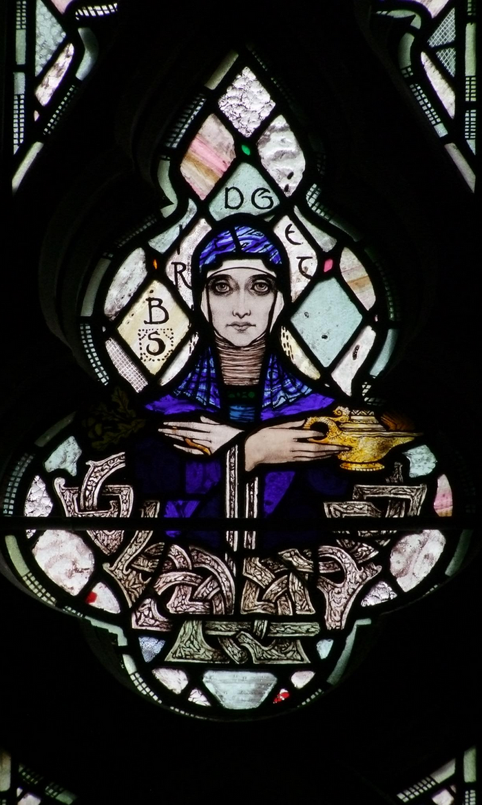Harry Clarke's 1918 stained-glass Brigid holds a lamp, recalling the flame tradition shared by the goddess and Saint Brigid.