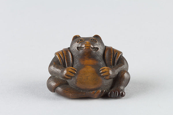 19th-century netsuke of a seated tanuki with a round belly, beating it in the manner of the hara tsutsumi legend.