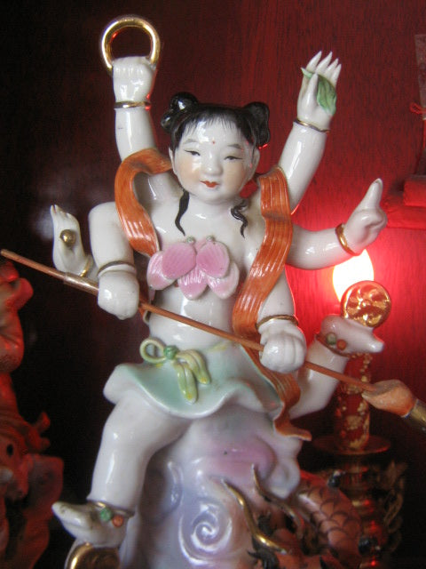 Porcelain figurine of Nezha with multiple arms, lotus-style bodice and sash, holding a ring and spear, riding on a dragon.