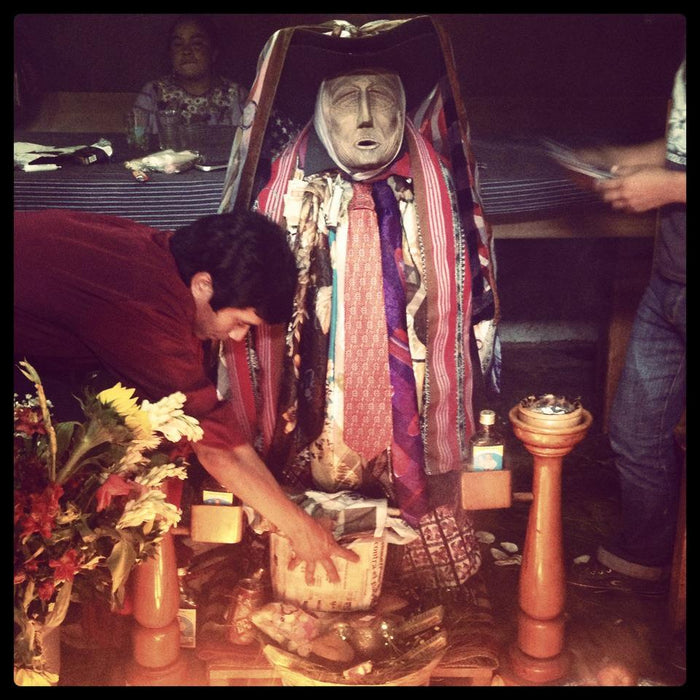Rilaj Mam with carved mask, hat, neckties and scarves; offerings and an incense stand surround the figure.