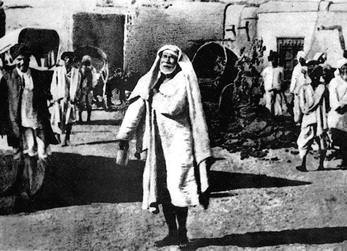 Sai Baba walking through Shirdi village, c. 1910, in a long white robe and head-cloth, with villagers behind.